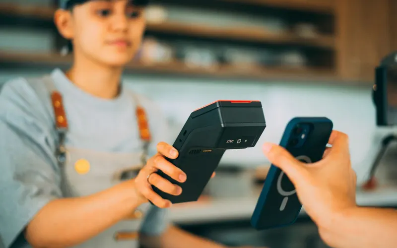 A member of counter staff in an apron lifts a handheld scanner up to the phone a customer is holding across the counter.